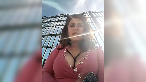 Media: A video of a plus-sized woman with large breasts, wearing a pink floral-patterned top, standing in front of a metal cage. Sunlight creates lens flare.