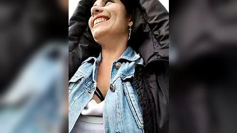 Media: Video of a smiling, light-skinned woman with short, dark hair, wearing a denim jacket over a white top, lying on a black jacket. Background is out of focus, suggesting outdoor setting.