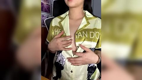 Media: Video of an Asian woman with medium skin tone, wearing a yellow-green floral button-up shirt, partially unbuttoned to reveal cleavage, hands placed on chest, blurred background.