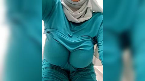 Media: Video of a woman in teal scrubs with a gray headscarf, hands on her pregnant belly, in a blurred teal background, likely a hospital or clinic setting.