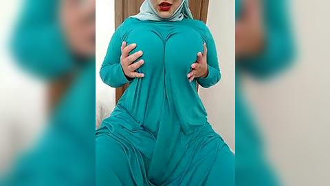 Media: Video of a woman in a teal, long-sleeved, form-fitting jumpsuit, sitting with legs spread. Her hands are cupping her large, round breasts. Background includes a beige wall and a wooden door.