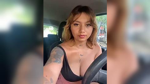 Media: Video of a young Asian woman with shoulder-length wavy hair, wearing a low-cut black top, driving a car, with blurry background and green trees outside.