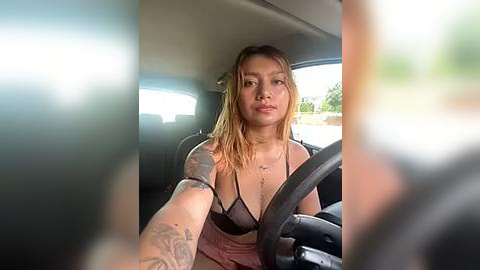 Media: Video of a young woman with medium skin tone, straightened blonde hair, and a tattooed arm, wearing a black bra, driving a car in daylight.