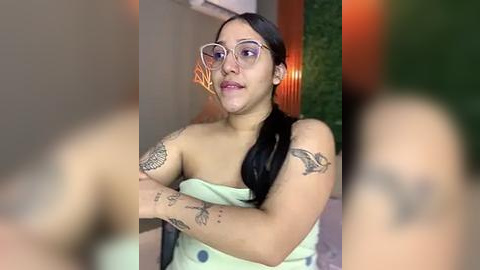 Media: Video of a Latina woman with medium skin tone, long black hair, wearing glasses and a light green strapless top, showcasing tattoos on her arms, sitting indoors with blurred background.