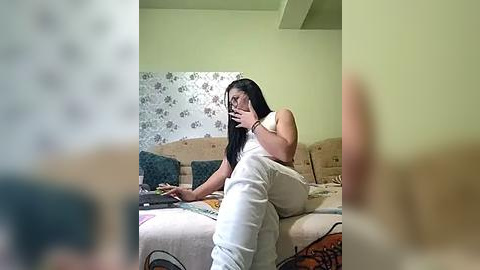 Media: A candid video captures a woman with long black hair, wearing a white shirt and pants, seated on a beige sofa with floral wallpaper. She is holding a phone to her ear, appearing engrossed in conversation.