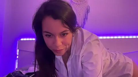 Media: Video of a light-skinned woman with long dark hair, wearing a white shirt, leaning forward on a bed with purple LED lighting, smiling seductively.