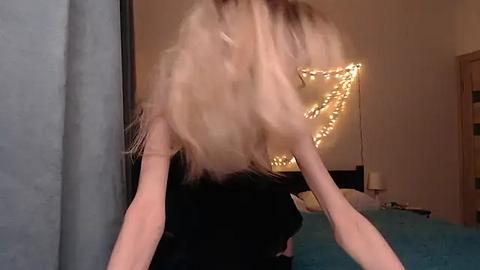 Media: Video of a blonde woman with long hair, captured in mid-motion, being pulled by hands from behind. Background features a teal bedspread, a warm string light display, and a wooden door.