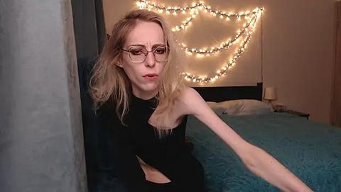 Media: Video of a slender, fair-skinned woman with blonde hair, wearing glasses and a black top, leaning against a wall in a dimly lit bedroom adorned with teal bedding and warm, string fairy lights.