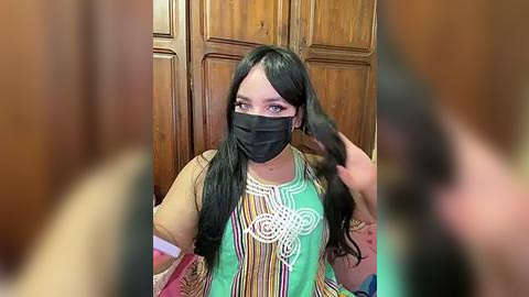 Media: Video of a young woman with long black hair, wearing a black face mask, a green blouse with white embroidery, and a black glove, sitting indoors with wooden paneling in the background.