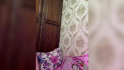 Media: Video of a cozy bedroom corner with a dark wooden door, floral wallpaper, and a bed with pink sheets adorned with cartoon characters, creating a playful and vintage aesthetic.