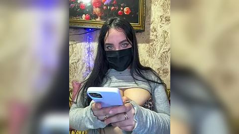 Media: Video of a woman with long dark hair, wearing a gray sweater, black face mask, and a phone, exposing her left breast. Background includes a framed painting and a textured wall.