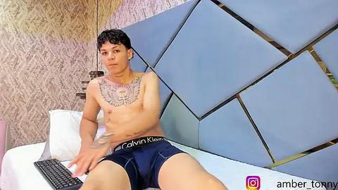 Media: Video of a shirtless, tattooed Asian man in Calvin Klein boxers, typing on a laptop on a bed with a geometric, light blue headboard. Background features floral-patterned wallpaper.