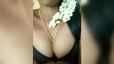 Media: A close-up video of a woman with medium brown skin wearing a black lace bra, adorned with a gold chain necklace featuring a large white flower pendant.