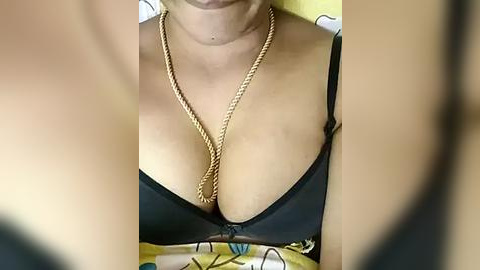 Media: A close-up video of a woman's cleavage, showing a black bra with a gold chain necklace. The background is blurred, with a yellow and black pattern visible.