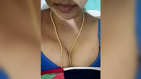 Media: Video of a woman's cleavage, wearing a blue bikini top and a gold chain necklace. Her face is partially visible with a slight smile, and her skin tone is light. The background is blurred, featuring a teal wall.