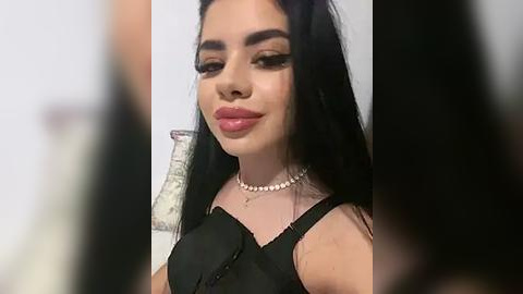 Media: A close-up video of a Latina woman with long, straight black hair, wearing a black sleeveless top, a pearl necklace, and bold makeup.