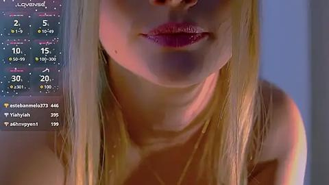 Media: Video of a young woman with fair skin, long blonde hair, and full lips. She is partially nude, with a soft focus background. A digital clock with current weather and temperature is visible in the top left corner.