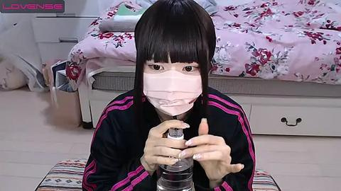 Media: Video of an Asian woman with straight black hair and pale skin, wearing a pink face mask and black athletic jacket with pink stripes, kneeling on a striped cushion, pouring water from a clear bottle into a glass.