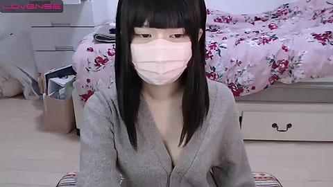 Media: A video of an Asian woman with straight black hair and a mask, wearing a gray cardigan, kneeling on a beige carpet in a bedroom with floral-patterned bedding and white furniture.