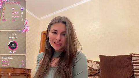 Media: Video of a young woman with long brown hair, fair skin, wearing a light green top, sitting in a cozy, beige-walled room with floral wallpaper, and a wooden table in the background.