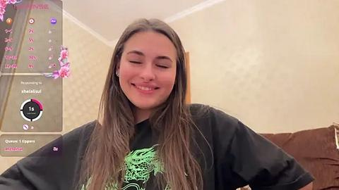 Media: Video of a smiling young woman with long, straight brown hair, wearing a black graphic T-shirt, standing in a dimly lit room with floral wallpaper and a digital clock.