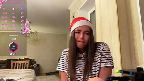 Media: Video of a young woman with long brown hair in a Santa hat, smiling, wearing a striped shirt, indoors with a TV displaying health stats, a wooden chair, and a cluttered room in the background.