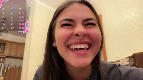 Media: Video of a smiling young woman with long brown hair, wearing a dark top, in a cozy kitchen with wooden cabinets and floral wallpaper.