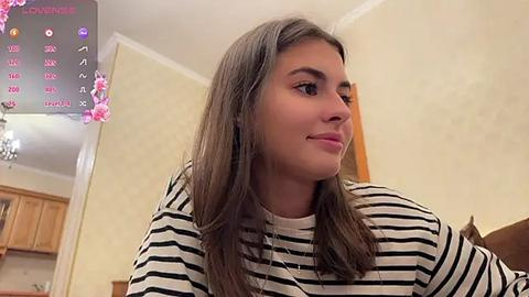 Media: Video of a young woman with light brown hair and fair skin, wearing a black and white striped top, sitting in a warmly lit, beige-walled room with a pink floral backdrop.