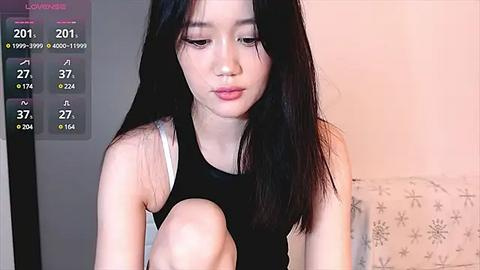 Media: A video of an Asian woman with long black hair, wearing a black tank top, sitting on a bed with a floral-patterned sheet.