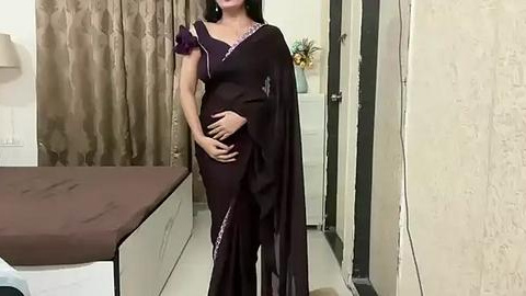 Media: Video of a pregnant woman in a dark purple saree, standing in a modern, beige-tiled bedroom with a bed, curtains, and a white dresser in the background.
