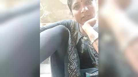 Media: A video of a South Asian woman with medium skin tone, short black hair, and traditional attire featuring a black embroidered blouse and matching shawl. She has a thoughtful expression, sitting outdoors, with blurred background.