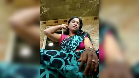 Media: Video of a South Asian woman with medium skin tone and long black hair, wearing a bright pink blouse and blue sari with geometric patterns, resting her head on her hand in a rustic, possibly rural, setting.