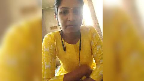 Media: Video of a young South Asian woman with short black hair, wearing a yellow kurta with white floral pattern, sitting with hands clasped, slightly blurred in the foreground.