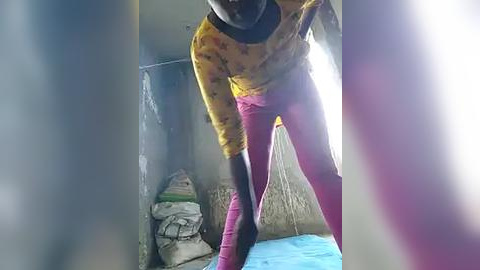 Media: Video of a person with dark skin, wearing a yellow shirt with black sleeves, purple pants, and a blue towel, bending over in a dimly lit, cluttered room with a bed and stacked clothes.