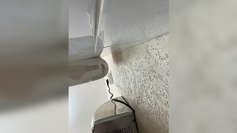 Media: A video of a white ceiling with a metallic air vent and a black power cord, capturing a close-up of a corner in a room. The background is slightly blurred, emphasizing the textured ceiling and the vent.