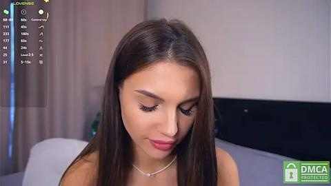 Media: A video of a young woman with long brown hair and light skin, wearing a silver necklace, smiling slightly, in a modern bedroom with a black TV and white curtains.