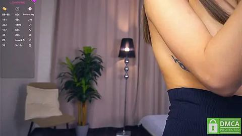 Media: A video of a woman's arm in a sleeveless top, with a tattoo visible. Background features a beige armchair, a tall floor lamp, and a potted plant.