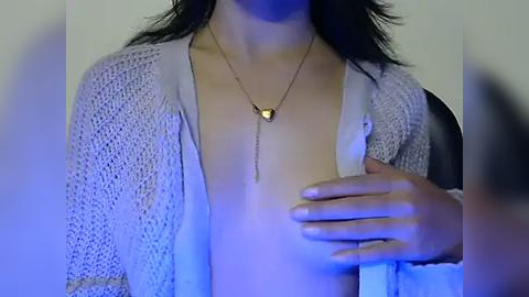 Media: Video of a light-skinned woman with dark hair, wearing a white, textured cardigan open to reveal a gold pendant necklace, and a hand on her chest. Background is blurred.