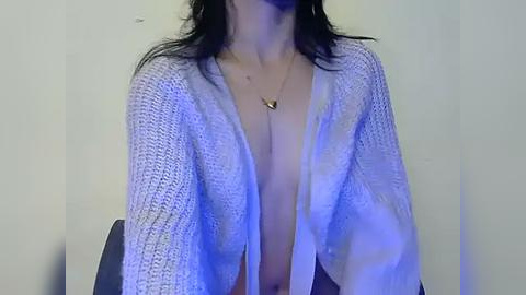 Media: Video of a topless woman with fair skin, wearing a loose, knit, white cardigan, revealing her medium-sized breasts. She has dark, wavy hair. Background is plain, off-white wall.