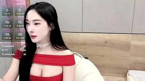 Media: Video of an Asian woman with long black hair, wearing a red off-shoulder dress, sitting on a white leather couch in a modern room.