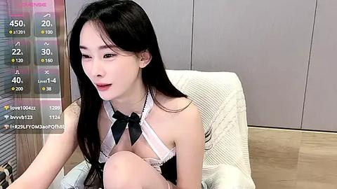 Media: Video of a slender, East Asian woman with long black hair, wearing a revealing white halter top with a black bow and black tie, sitting on a beige armchair in a modern room with grey cabinets and wooden flooring.