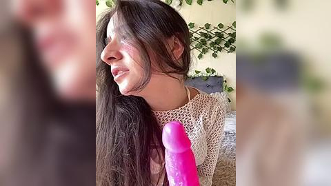 Media: Video of a young woman with long, straight brown hair, wearing a beige crochet top, holding a large pink dildo. The background features a beige wall with green vines.