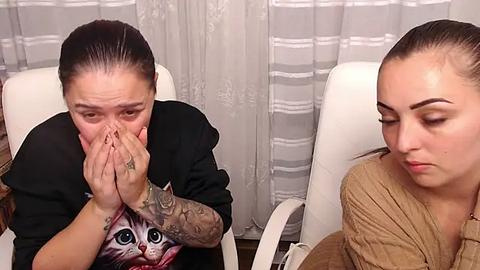 Media: Video of two women with light skin tones, one with a tattooed arm covering her face, the other with a tan sweater and long hair, sitting on white chairs in a room with striped curtains.