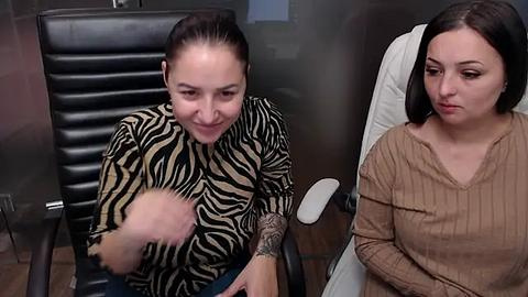 Media: Video of two women in a medical setting. One, with light skin and dark hair, wears a zebra-print top, tenderly touches a bald patient's head. The other, with dark hair and fair skin, wears a beige sweater, sits nearby, appearing concerned.