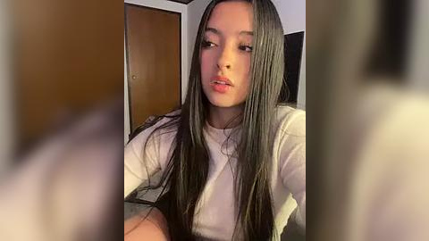 Media: Video of a young Asian woman with straight, long black hair, wearing a white t-shirt, leaning forward with a neutral expression, indoors with a wooden door and blurred background.