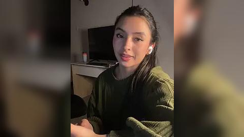 Media: Video of a young Asian woman with straight black hair, wearing a green sweater, sitting in a dimly lit room with blurred figures in the background.