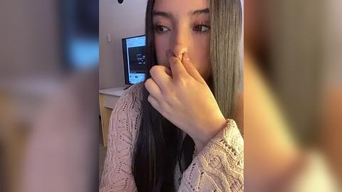 Media: Video of a young woman with long, straight black hair, wearing a beige crochet sweater, covering her mouth with her right hand, in a dimly lit room with a computer screen visible in the background.