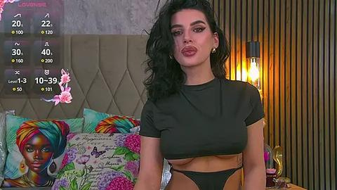 Media: Video of a curvy woman with dark hair, wearing a black crop top and high-waisted pants, sitting on a bed with colorful pillows, in a modern bedroom with a headboard and lamp.