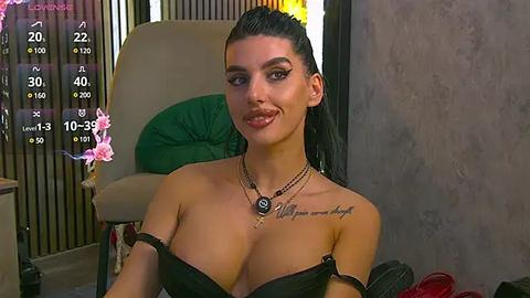 Media: Video of a young woman with long black hair styled in a high ponytail, wearing black lingerie, sitting in a modern room with a green chair, floral decorations, and a textured wall.