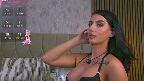 Media: Video of a woman with long black hair and tanned skin, wearing a black bra, in a bedroom with a beige quilted headboard and floral wallpaper.